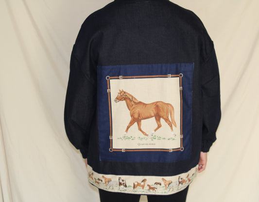 Handmade dark blue denim jacket featuring a large vintage horse illustration on the back and a hem trimmed with printed fabric showing a galloping horse motif. Western inspired tan accents and a denim panel across the front. One-of-a-kind ethically sewn from rescued textiles.