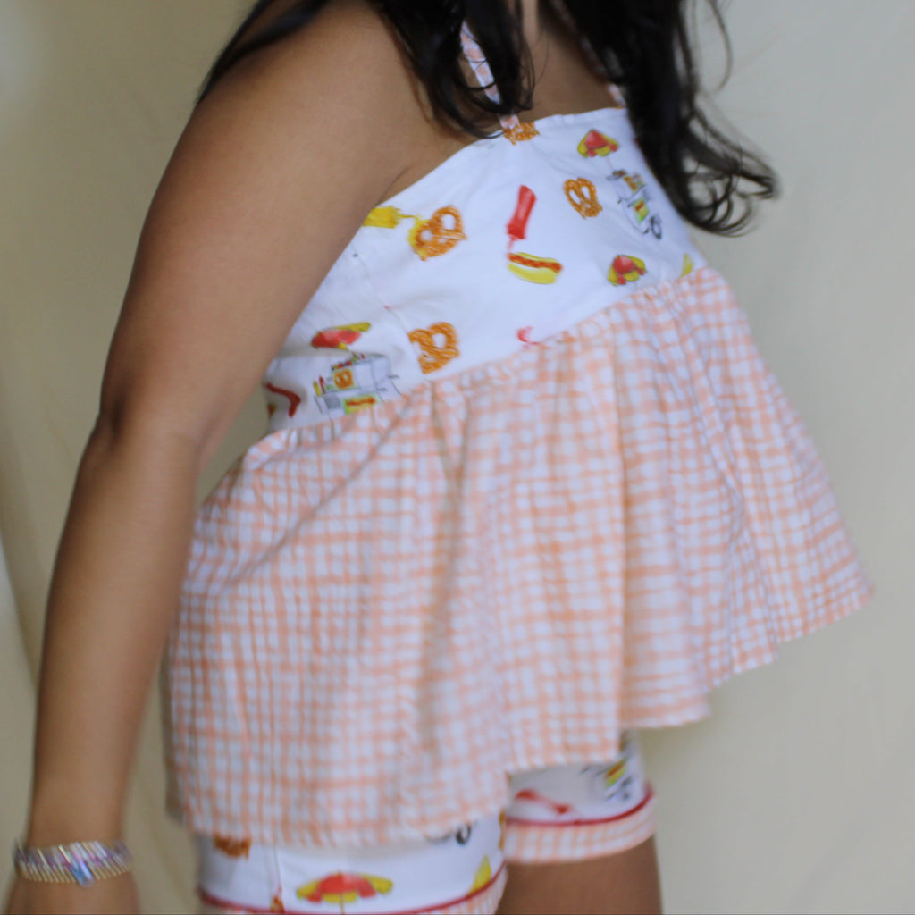 Woman wearing a peach gingham babydoll halter top with playful snack-print bust panel featuring hotdogs, pretzels, and condiments. Ethically handmade summer top from upcycled deadstock fabric, styled against a neutral backdrop. 