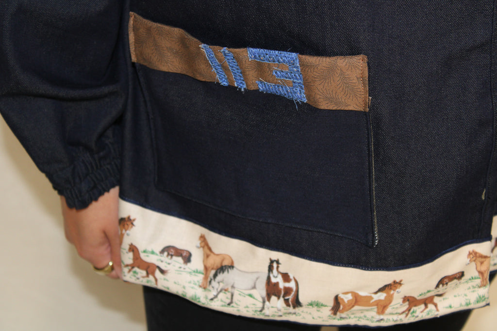Handmade dark blue denim jacket featuring a large vintage horse illustration on the back and a hem trimmed with printed fabric showing a galloping horse motif. Western inspired tan accents and a denim panel across the front. One-of-a-kind ethically sewn from rescued textiles. 