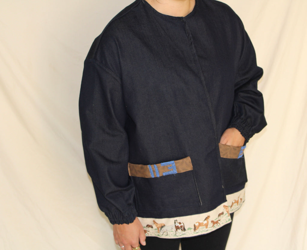 Handmade dark blue denim jacket featuring a large vintage horse illustration on the back and a hem trimmed with printed fabric showing a galloping horse motif. Western inspired tan accents and a denim panel across the front. One-of-a-kind ethically sewn from rescued textiles.